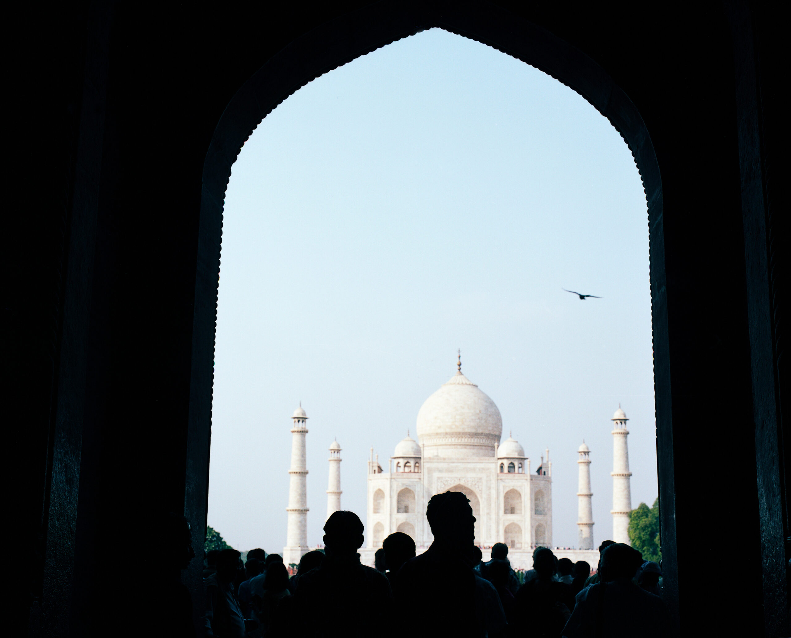 Taj Mahal im Gegenlicht – Blick durch das dunkle Eingangstor auf das leuchtende Mausoleum, mit Menschen im Vorhof Wärend einer Fotoreise in Indien: Frontaler Blick auf das Taj Mahal durch ein dunkles Eingangstor – der helle Marmorbau leuchtet im Kontrast zum Schatten des Portals. Menschen bewegen sich wie Sillhouetten durch den Vorhof. Ein Bild voller Tiefe, Lichtstimmung und ungewöhnlicher Perspektive auf das berühmte Monument.